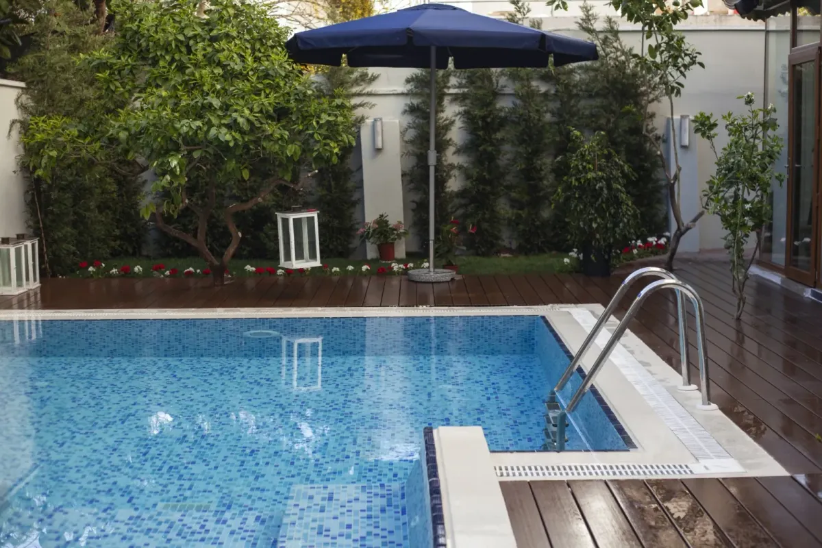 Piscina de gresite azul terminada en el jardín de un chalet, con escalera de acero, tarima de madera y sombrilla